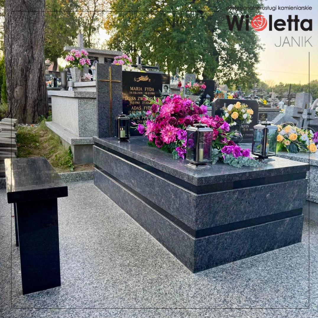 Granitowy nagrobek dwuosobowy w ciemnej tonacji, zdobiony kwiatami i lampionami, na tle cmentarza. Obok ławka z tego samego kamienia.