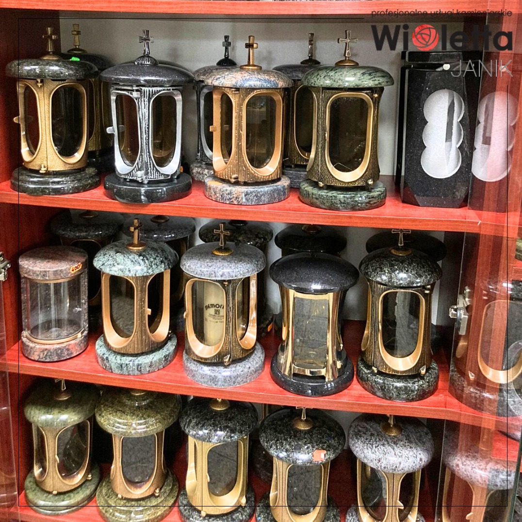 Różnorodne lampiony nagrobne z kamienia i metalu, prezentowane na czerwonych półkach. Widoczne różne wzory i kolory kamienia, z krzyżami na szczycie.