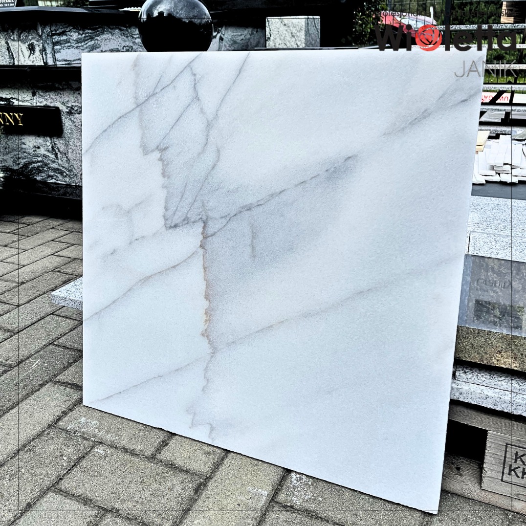 Płyta z jasnego marmuru o subtelnym użyleniu, ustawiona na brukowanej powierzchni. W tle widoczne inne płyty kamienne i elementy nagrobków.