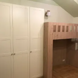 Biała szafa czterodrzwiowa z drewnianymi gałkami obok drewnianego łóżka na antresoli ze schodami w formie szafek, oświetlona kinkietem w pokoju z jasnymi ścianami i skośnym sufitem.