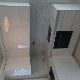 Nowoczesna, biała kuchnia z błyszczącymi frontami, marmurowym blatem i ścianą, widoczne otwory na gniazdka elektryczne w ścianie.