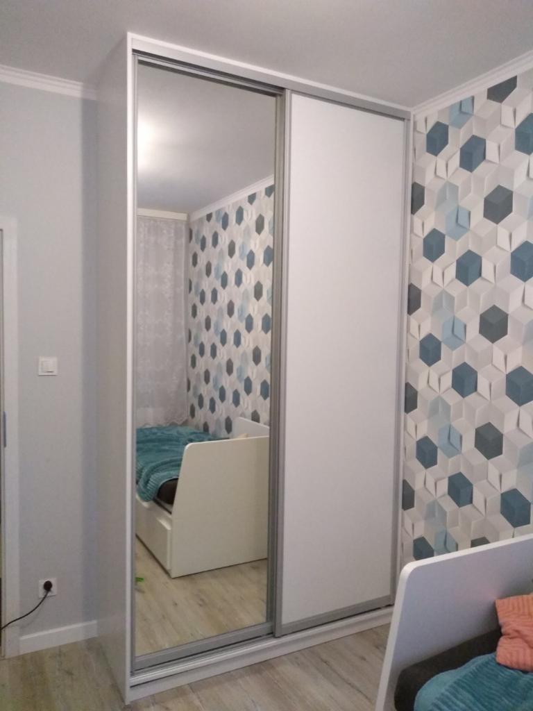 Biała szafa przesuwna z lustrem w pokoju dziecięcym, odbicie łóżka w lustrze, ściana z geometryczną tapetą.