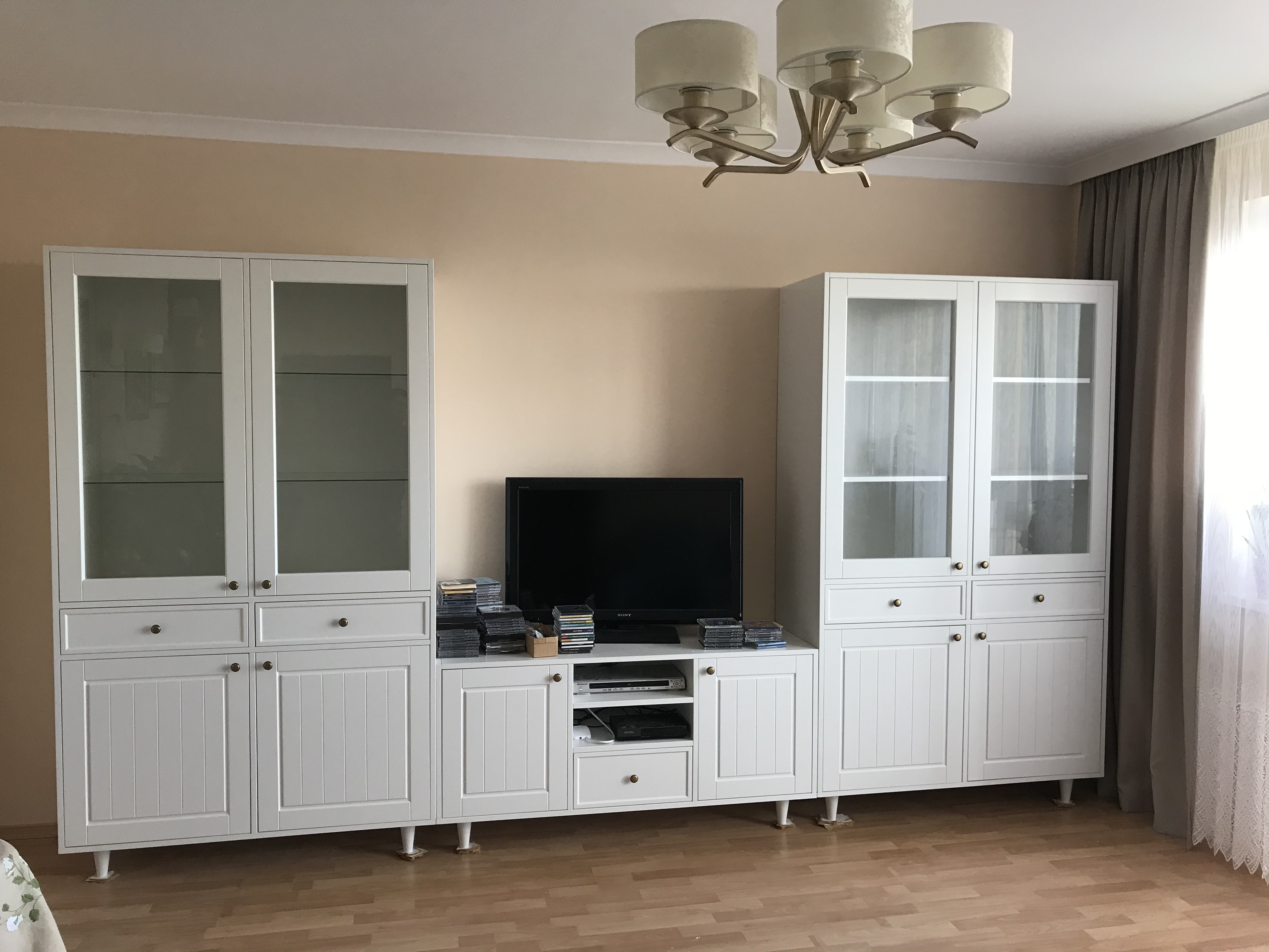 Biała, modułowa zabudowa ściany w salonie z elementami przeszklonymi, szafkami i szufladami, z telewizorem pośrodku i stertą płyt CD obok, całość oświetlona przez lampę sufitową z abażurami.