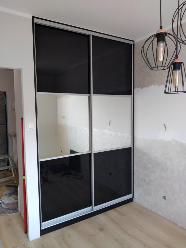 Czarno-biała przesuwna szafa z lustrzanymi panelami w trakcie montażu, widoczne niedokończone ściany, surowa żarówka i poziomica oparta o futrynę.
