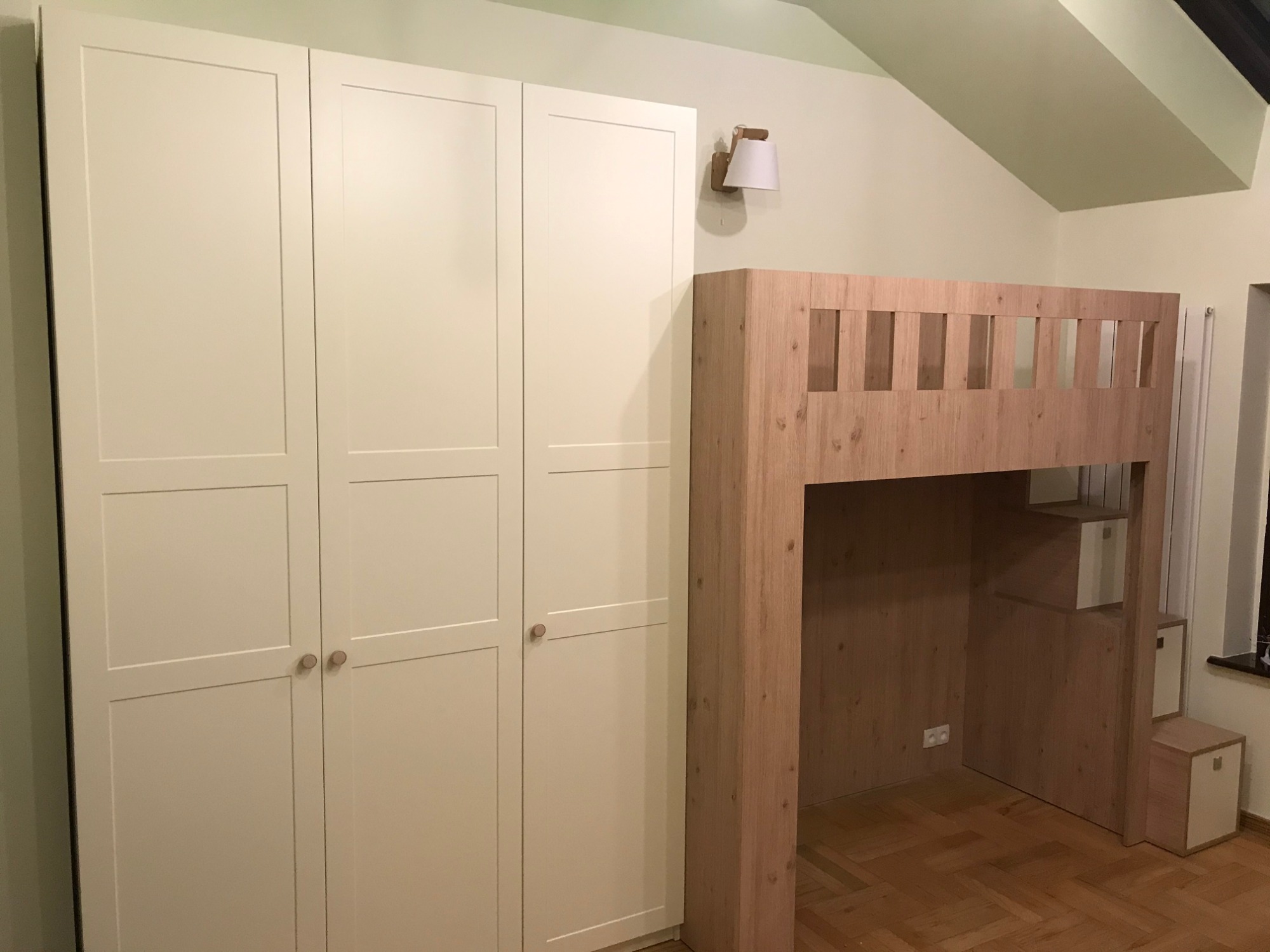 Biała szafa czterodrzwiowa z drewnianymi gałkami obok drewnianego łóżka na antresoli ze schodami w formie szafek, oświetlona kinkietem w pokoju z jasnymi ścianami i skośnym sufitem.
