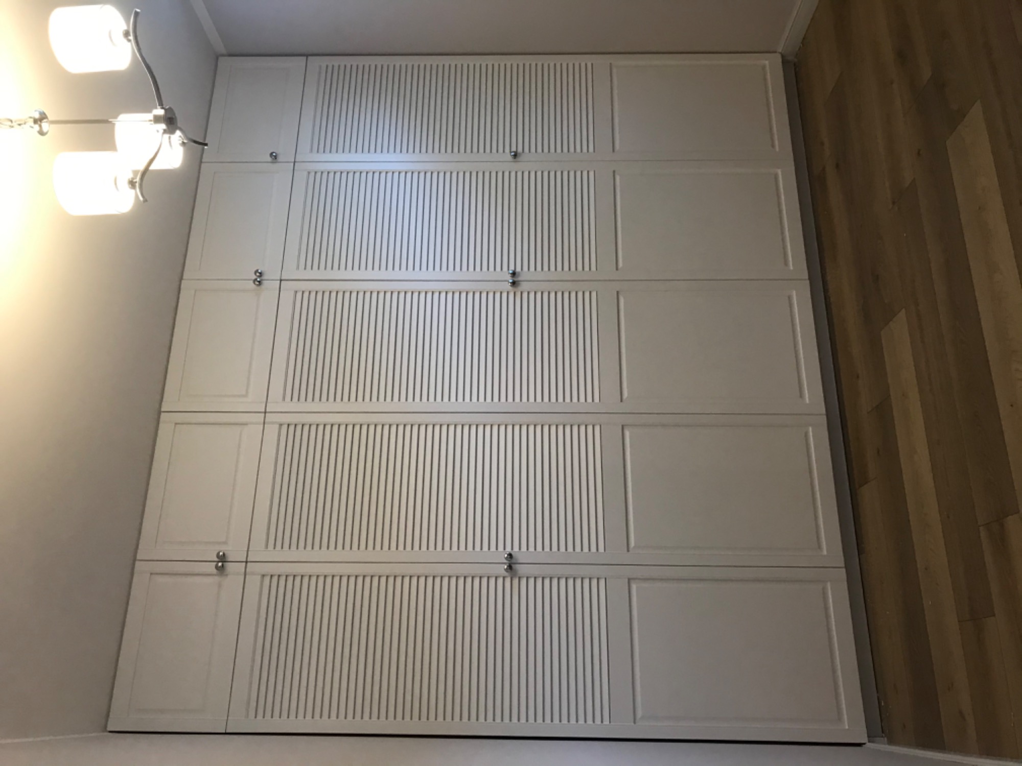 Wysoka, biała szafa wnękowa z lamelkowymi panelami na górnych drzwiach, widoczna lampa sufitowa i drewniana podłoga.