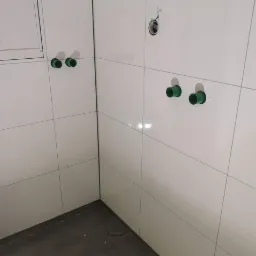 Białe, błyszczące płytki ścienne w rogu łazienki, z wystającymi zielonymi rurami instalacji wodnej i elektrycznej przed montażem armatury.