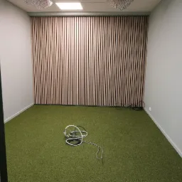 Wnętrze biura z wykończoną ścianą z drewnianych paneli pionowych, zielona wykładzina dywanowa, widoczne kable na podłodze.