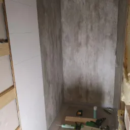 Wnętrze łazienki w trakcie remontu: panele ścienne imitujące beton i białe panele ułożone na ścianach, widoczne narzędzia i materiały budowlane na podłodze.