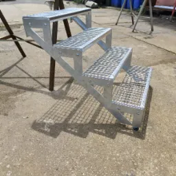 Zewnętrzne schody metalowe z kratownicowymi stopniami, ocynkowane, widoczne na betonowym podłożu, z wózkiem dostawczym i pojemnikami na śmieci w tle.