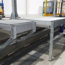 Stalowa platforma robocza z kratą pomostową, zamontowana na betonowej konstrukcji w hali przemysłowej, widoczny wózek paletowy i wiertarka udarowa w tle.