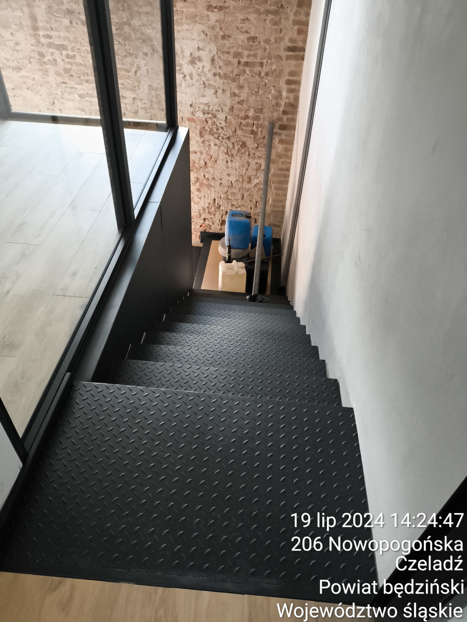 Metalowe schody z blachy ryflowanej w nowoczesnym wnętrzu z ceglaną ścianą w tle, widoczne narzędzia i pojemniki budowlane.