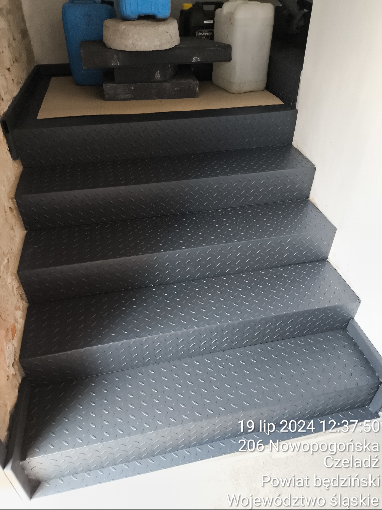 Metalowe schody z blachy ryflowanej, pomalowane na szaro, prowadzące do góry, z widocznymi śladami niedokończonego remontu na ścianie obok.