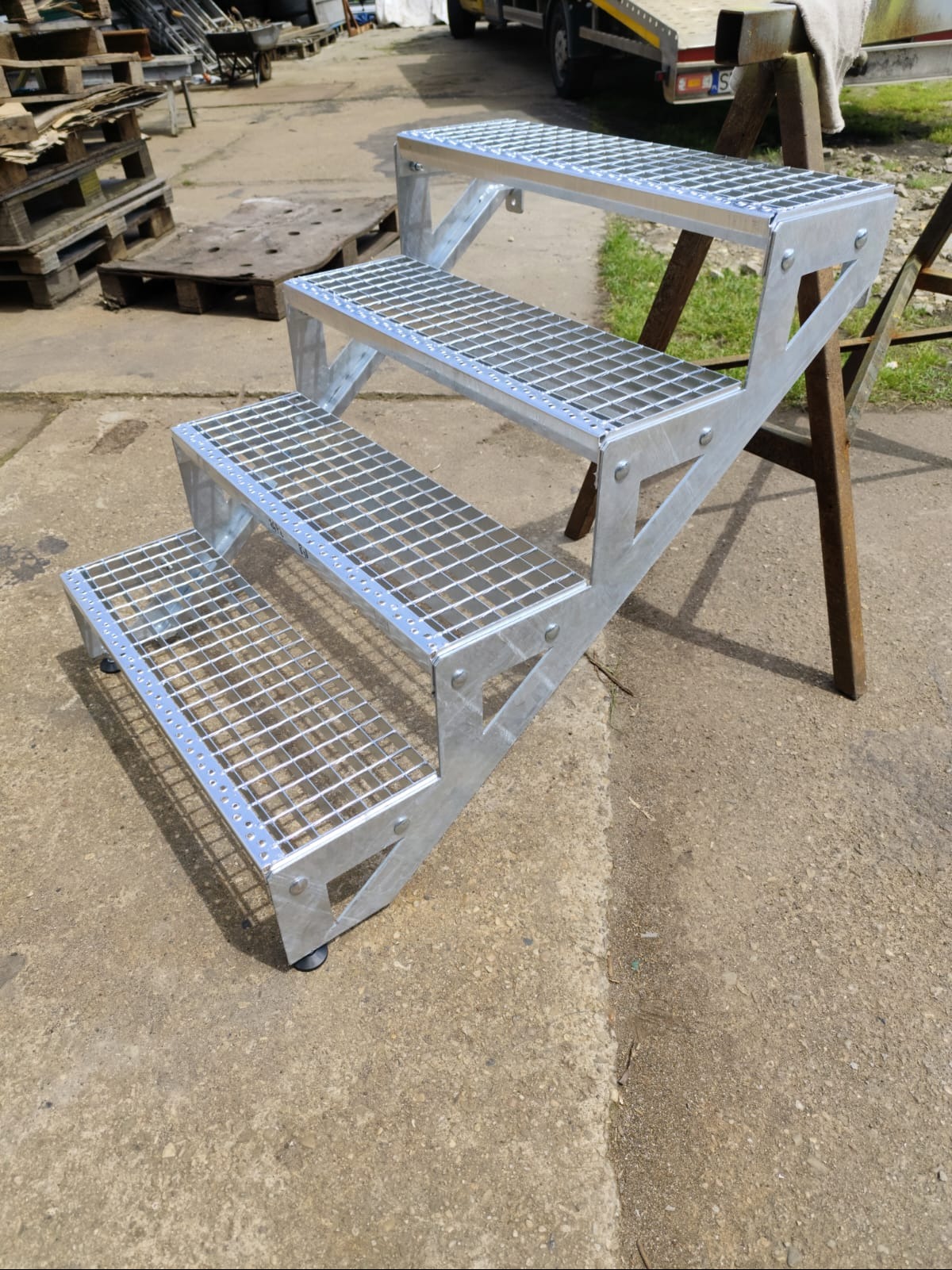 Trzy stopnie schodów z metalowej kraty, wykonane w Będzinie, ustawione na betonowym podłożu, widoczne nity i gumowe nóżki, w tle palety i przyczepa.