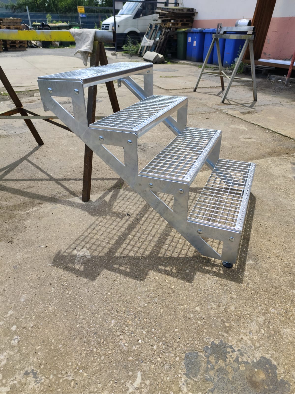 Zewnętrzne schody metalowe z kratownicowymi stopniami, ocynkowane, widoczne na betonowym podłożu, z wózkiem dostawczym i pojemnikami na śmieci w tle.