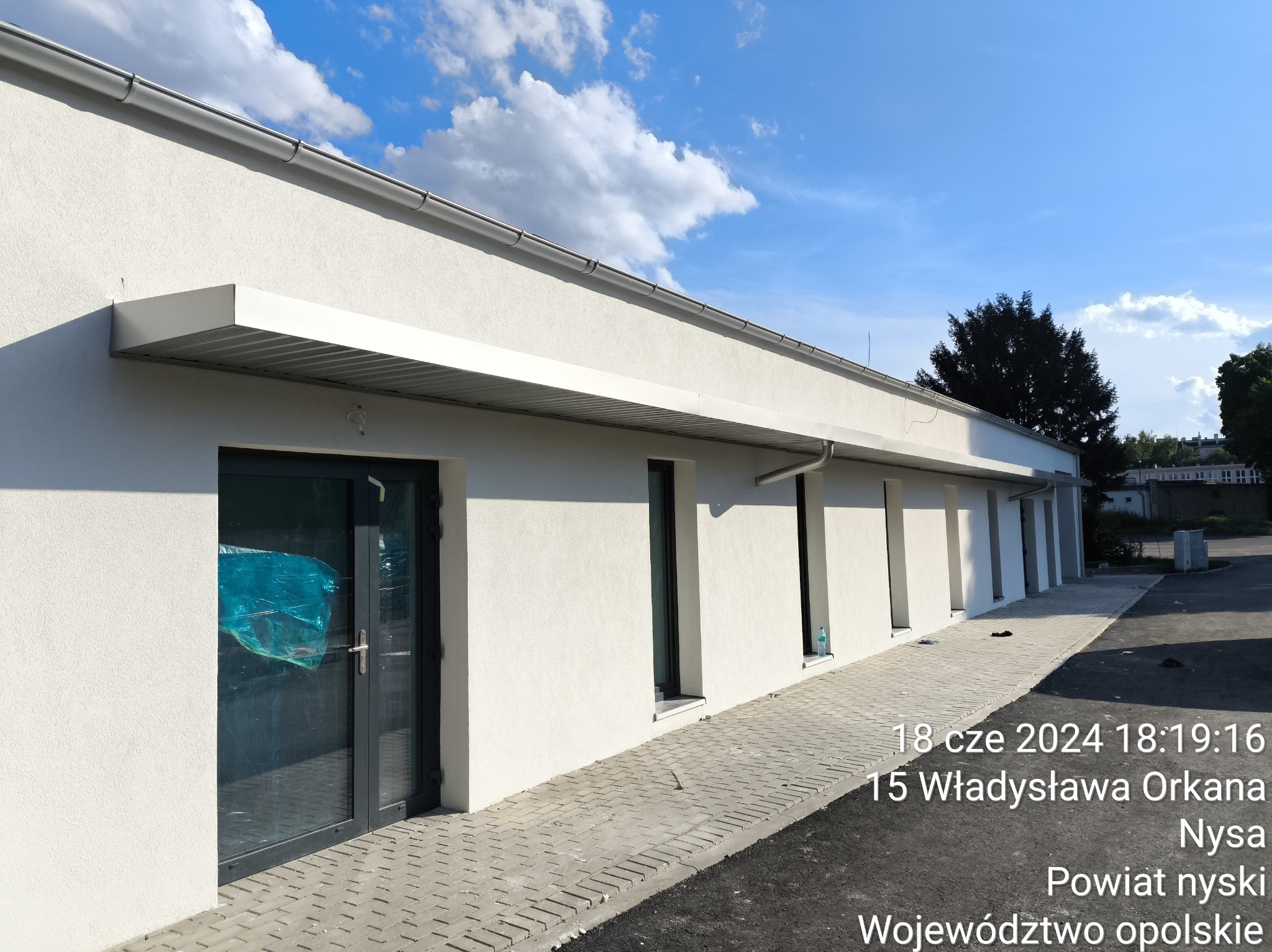 Biały budynek hali z szarymi ramami okiennymi i drzwiami, częściowo osłoniętymi folią ochronną, widok z zewnątrz w słoneczny dzień, z widocznym chodnikiem z kostki brukowej.