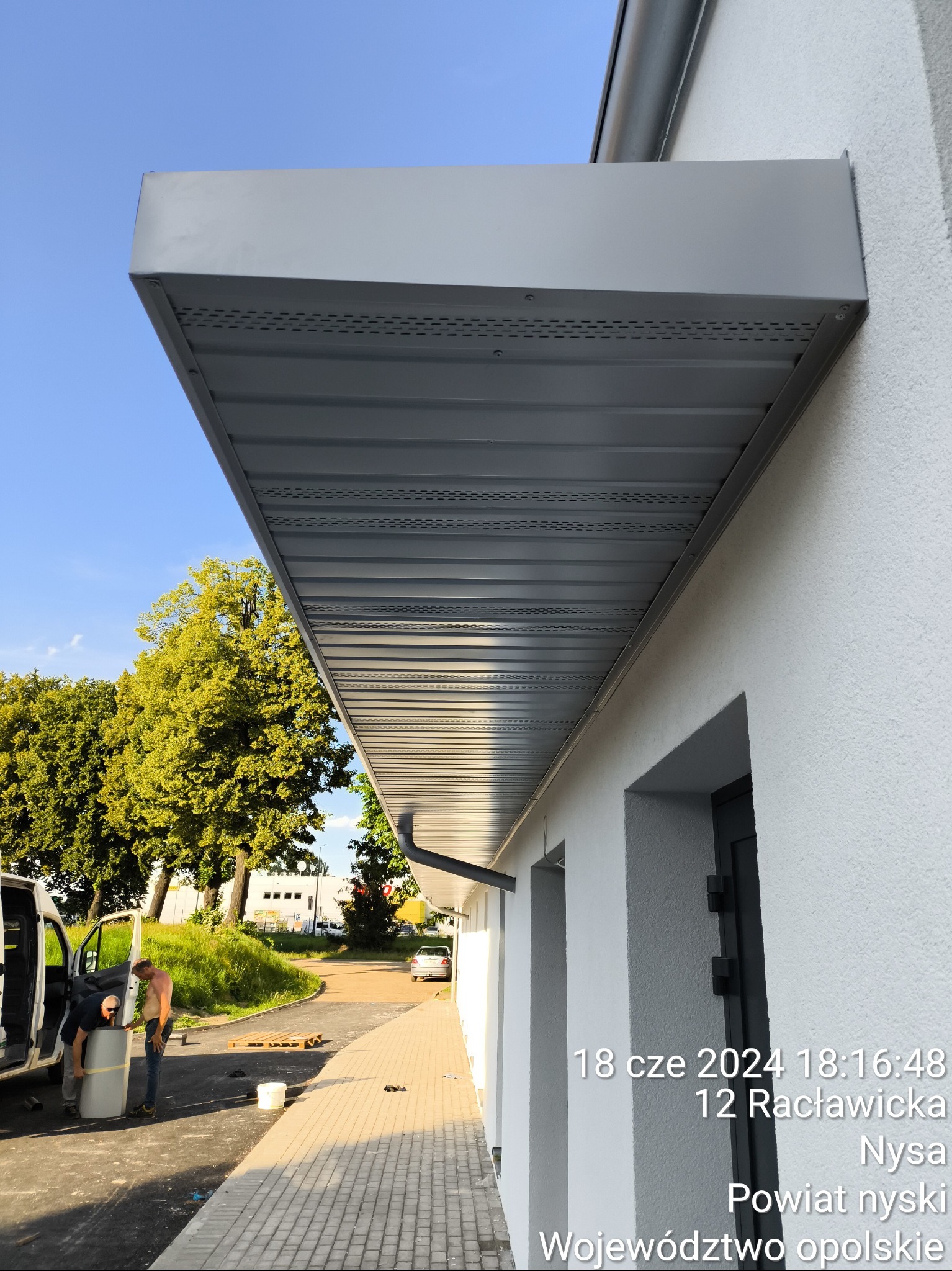 Montaż szarego zadaszenia nad wejściem do budynku z białym tynkiem, widoczni dwaj mężczyźni przygotowujący element konstrukcyjny przy samochodzie dostawczym.