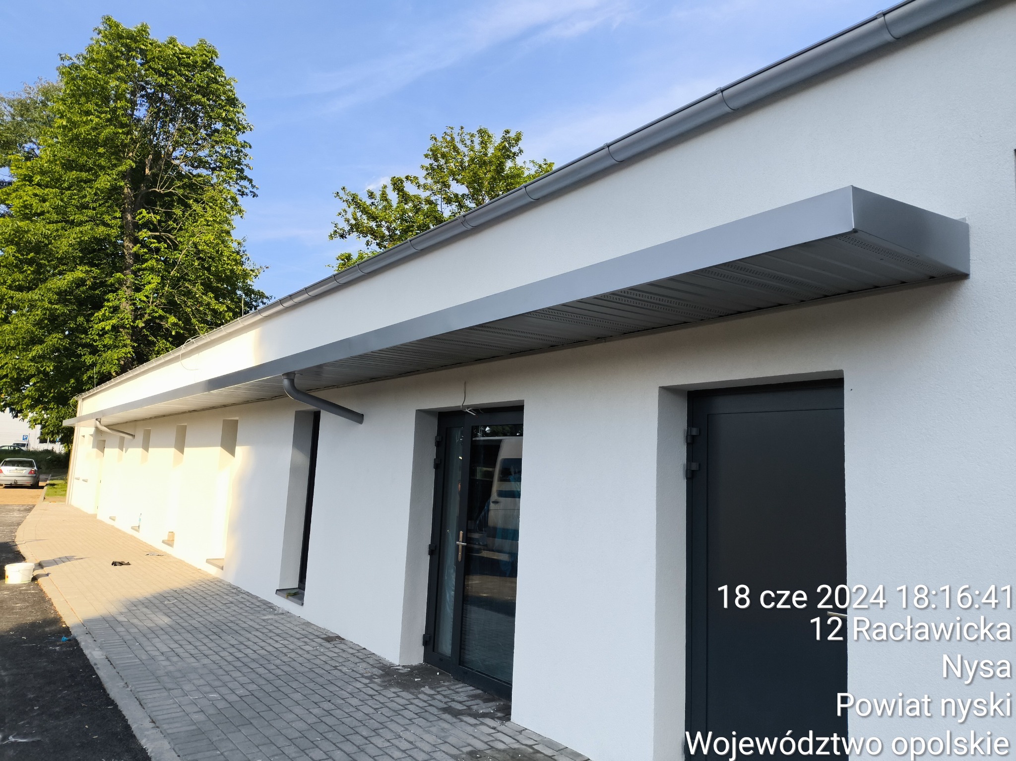 Nowoczesny budynek z białymi ścianami i szarymi, metalowymi daszkami nad wejściami, widoczny chodnik z kostki brukowej, drzewa w tle i niebieskie niebo, adres firmy widoczny na drzwiach.