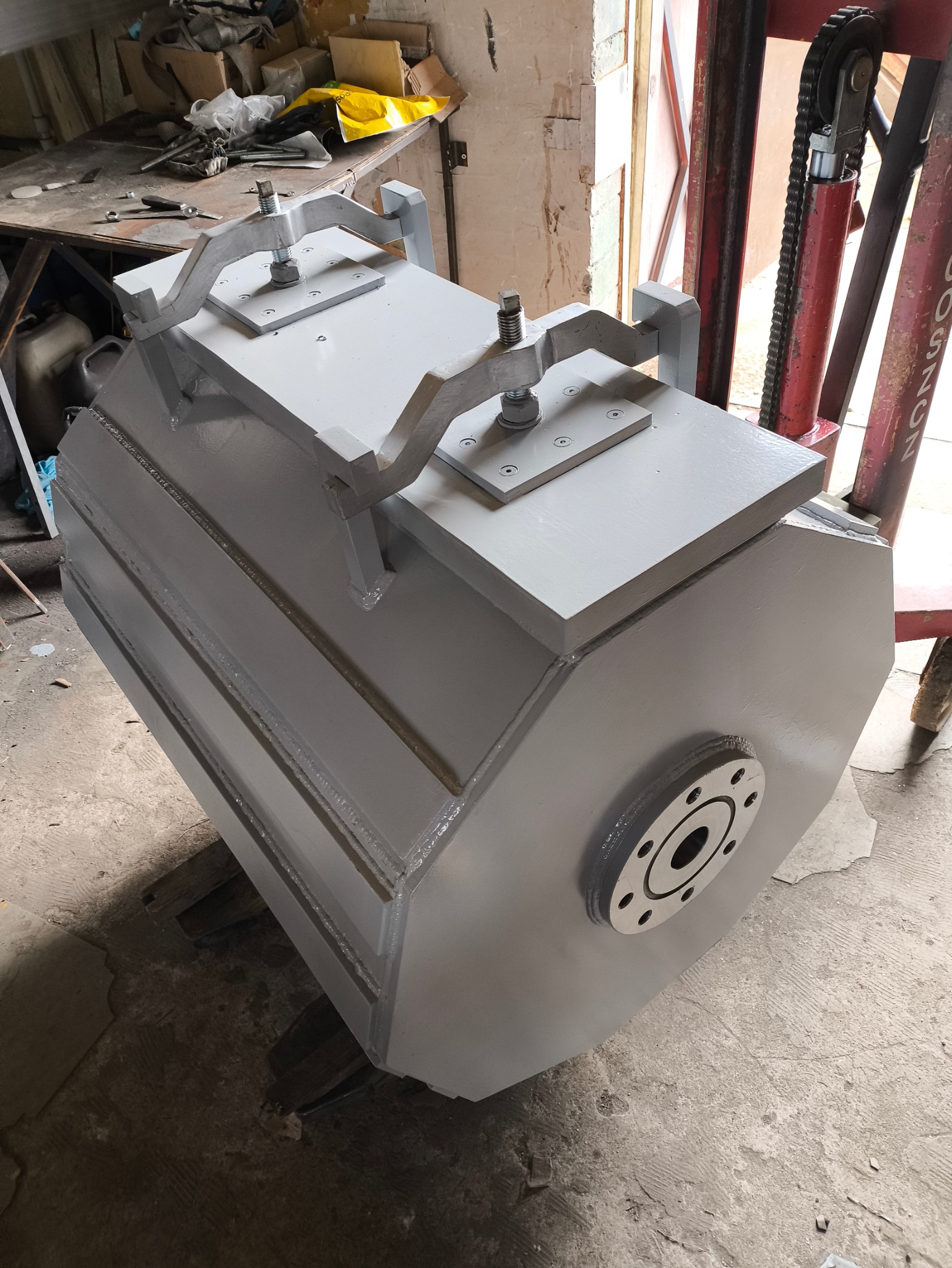 Szary, spawany walec z metalu z przymocowanymi uchwytami, umieszczony na palecie widłowej w warsztacie.