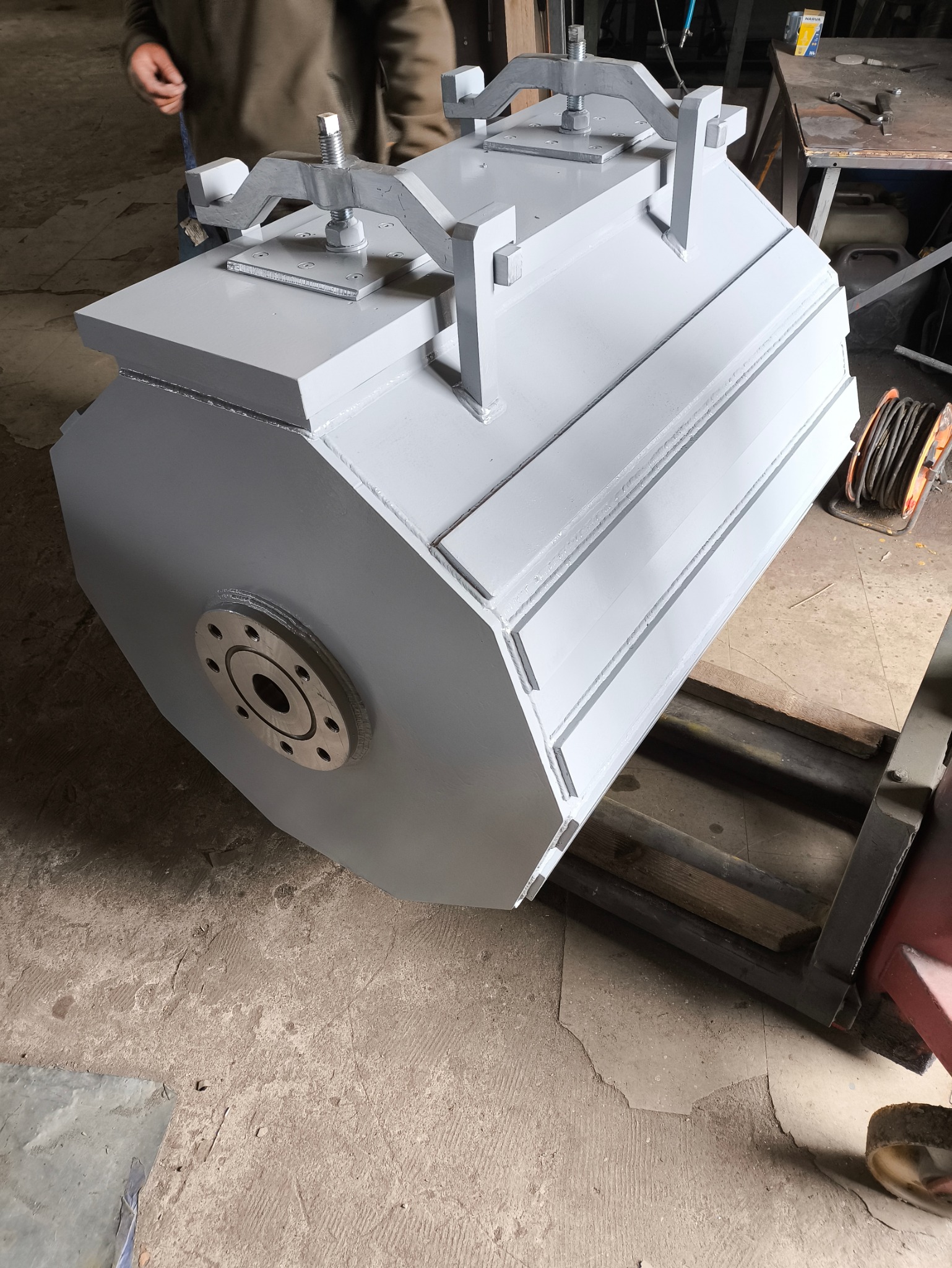 Szary, cylindryczny element konstrukcyjny z metalu z przyspawanymi uchwytami, widoczny na betonowej podłodze warsztatu.