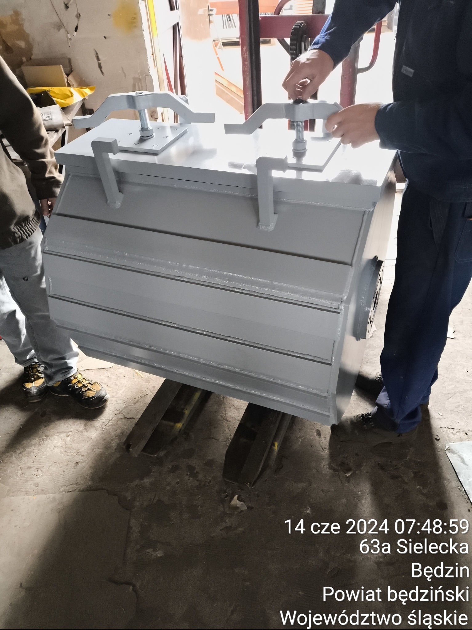 Szara, pomalowana konstrukcja stalowa z elementami mocującymi, podnoszona wózkiem widłowym w hali produkcyjnej, widoczne ręce pracownika regulujące mocowania.