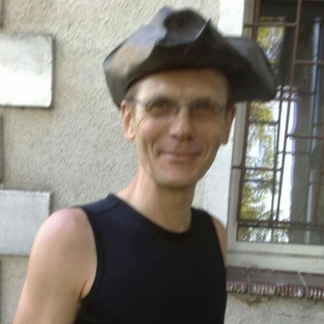 Portret uśmiechniętego mężczyzny w czarnej koszulce bez rękawów i kapeluszu pirackim, stojącego na zewnątrz na tle ściany z oknem.