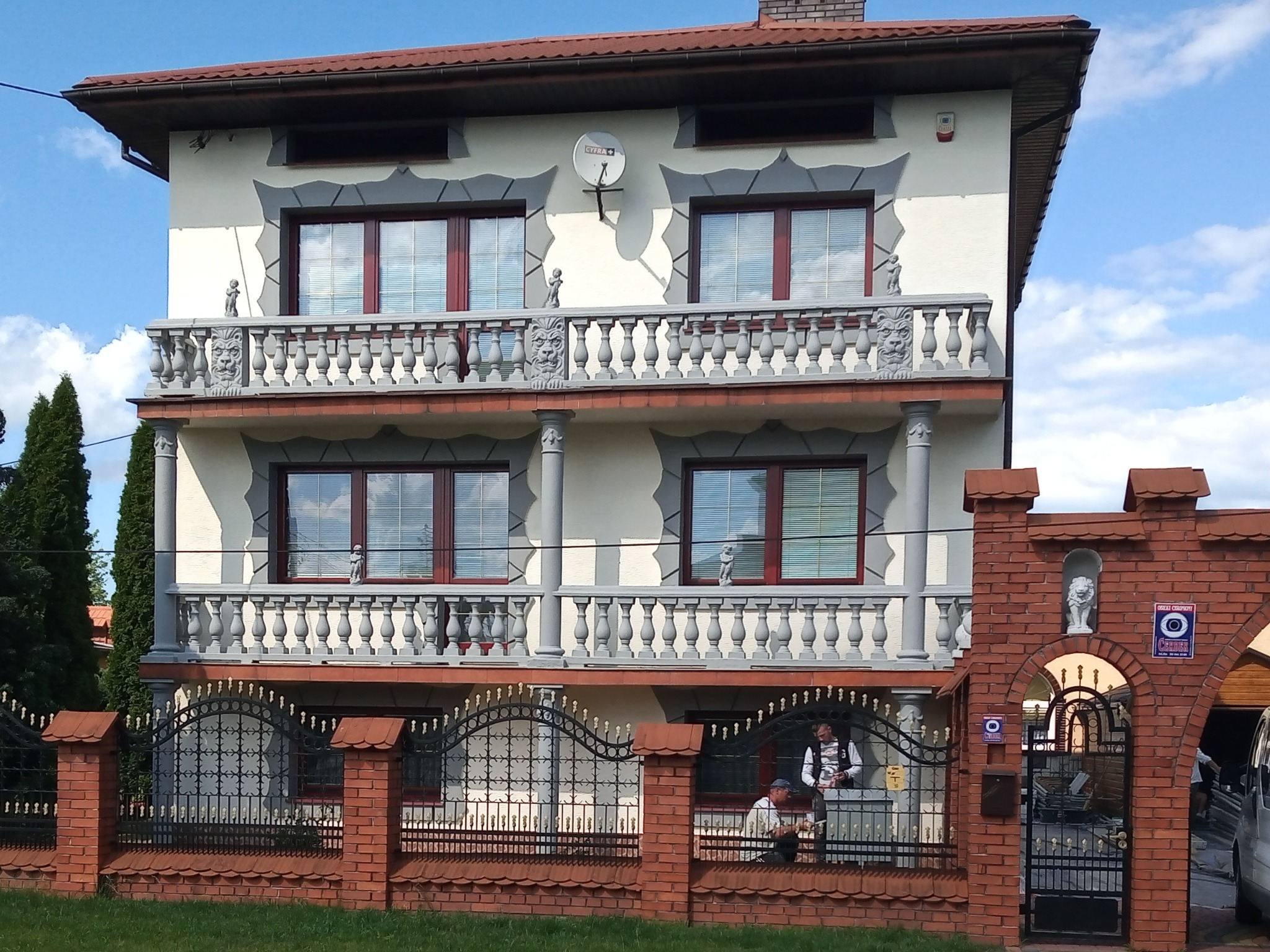 Dwupiętrowy dom z balkonami ozdobionymi rzeźbami lwów i figurkami, widoczne elementy dekoracyjne elewacji, brama wjazdowa z cegły z motywem lwa i pracownicy w tle.