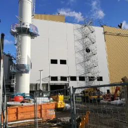 Budowa hali przemysłowej z widocznym kominem i rusztowaniami, sprzęt budowlany na placu budowy, częściowo ogrodzony teren.