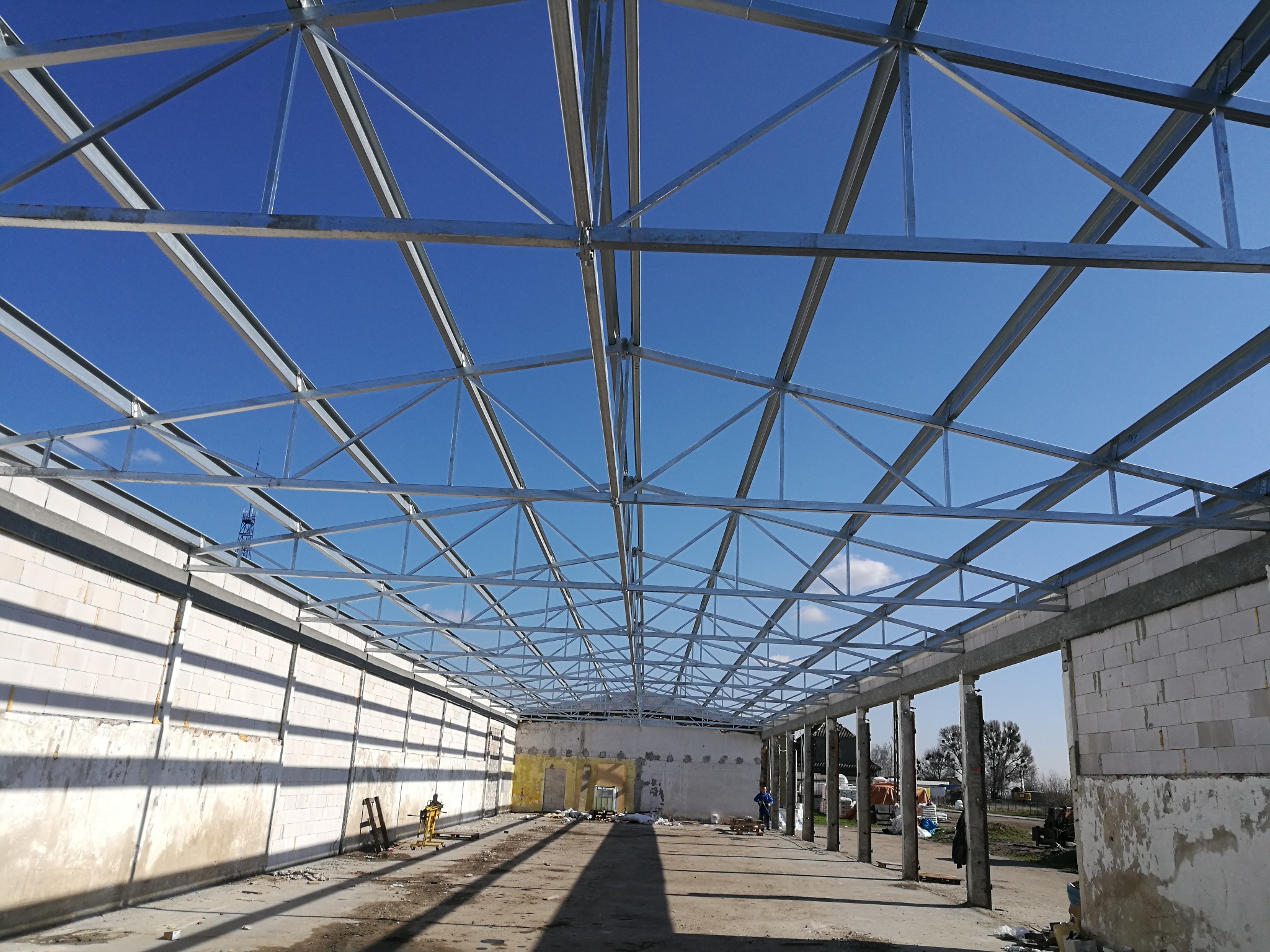 Wnętrze budowanej hali z widoczną stalową konstrukcją dachu, ściany z bloczków betonowych, podłoże w stanie surowym, błękitne niebo w tle.