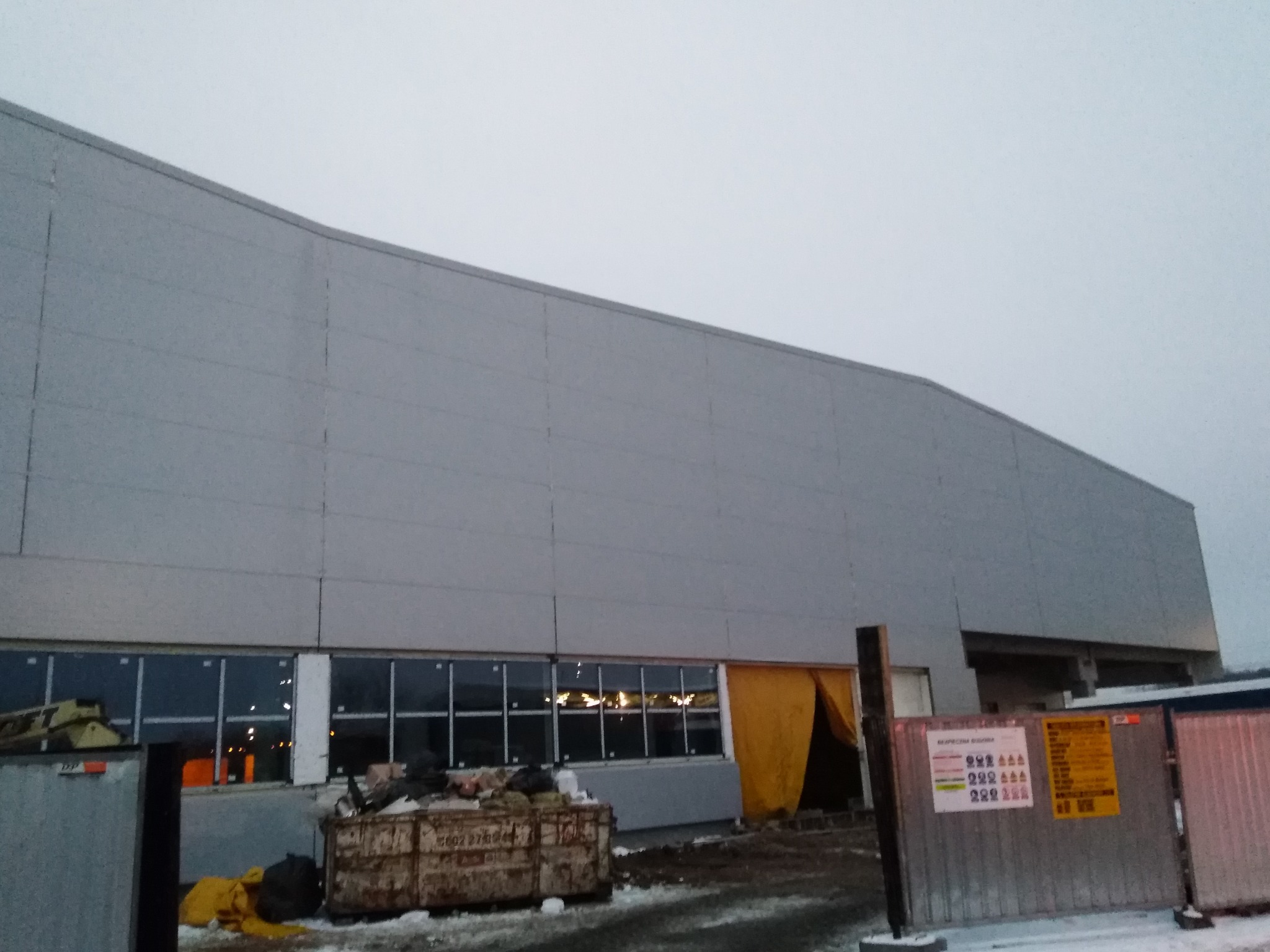 Budowa hali z szarej płyty warstwowej, widoczne okna i żółta plandeka w otworze wejściowym, kontener na odpady budowlane na pierwszym planie.