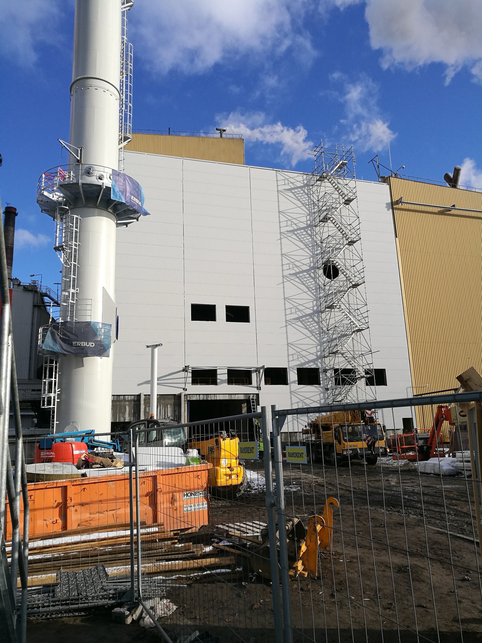 Budowa hali przemysłowej z widocznym kominem i rusztowaniami, sprzęt budowlany na placu budowy, częściowo ogrodzony teren.