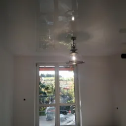 Biały, błyszczący sufit napinany z odbiciem okna i lampy w Smolnikach, widok na świeżo pomalowane ściany i drzwi balkonowe z widokiem na ogród.