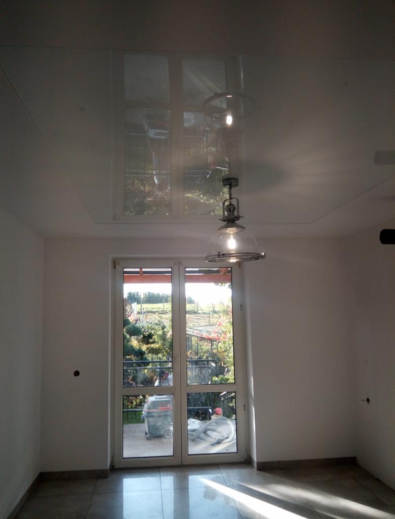 Biały, błyszczący sufit napinany z odbiciem okna i lampy w Smolnikach, widok na świeżo pomalowane ściany i drzwi balkonowe z widokiem na ogród.