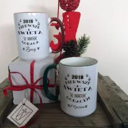 Dwa białe kubki z okolicznościowym nadrukiem '2018 Pierwsze Święta z Moją Gorącą Żoną/Mężem' na drewnianej skrzynce, obok prezent zawiązany czerwoną wstążką i świąteczna dekoracja.