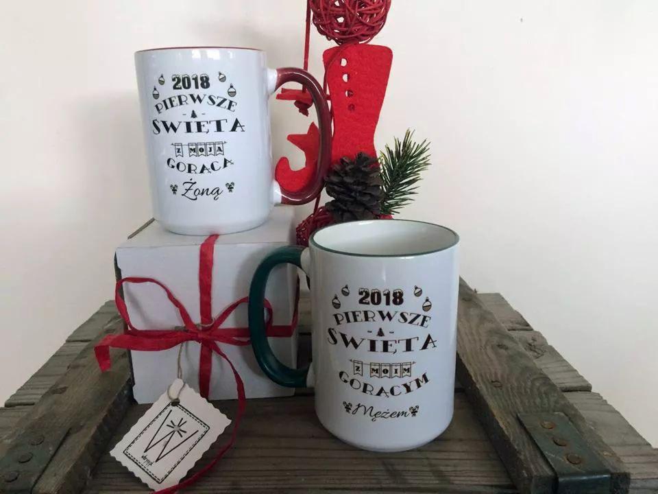 Dwa białe kubki z okolicznościowym nadrukiem '2018 Pierwsze Święta z Moją Gorącą Żoną/Mężem' na drewnianej skrzynce, obok prezent zawiązany czerwoną wstążką i świąteczna dekoracja.