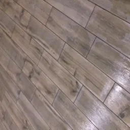 Ukośne ułożenie podłogowych płytek ceramicznych imitujących drewno, z widocznymi fugami i fragmentem ściany z listwą przypodłogową oraz wzorzystym dywanem w rogu.