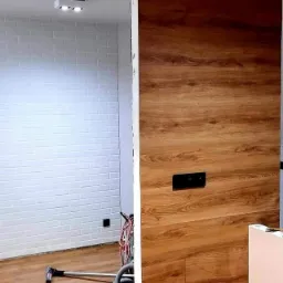 Ściana z paneli drewnopodobnych z gniazdkiem elektrycznym w trakcie montażu w Białymstoku, widoczny odkurzacz i biała ściana z cegiełek.