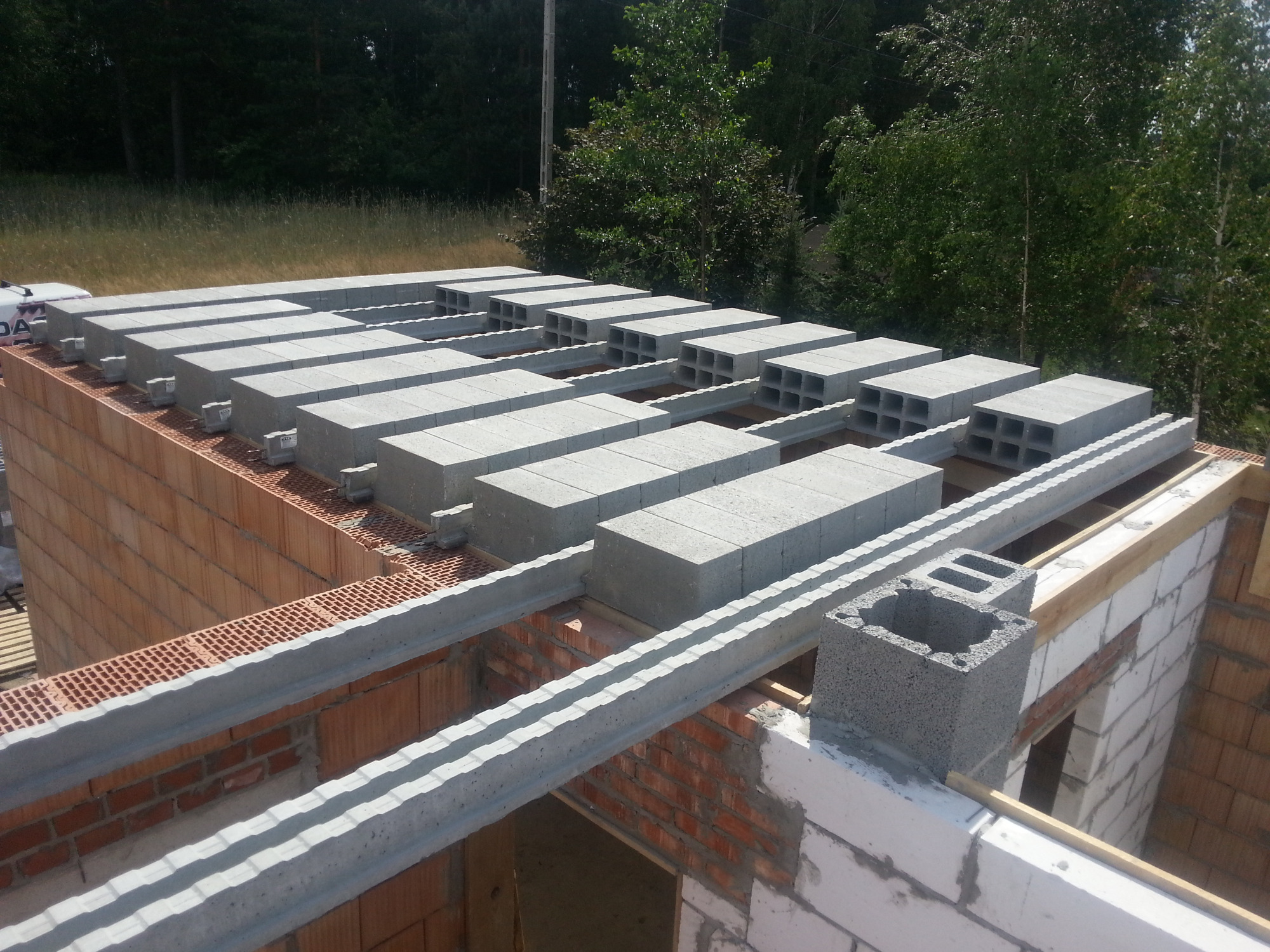 Wieniec stropowy z prefabrykatów betonowych na murach z cegły kratówki, widok z góry na budowę domu jednorodzinnego, w tle drzewa.