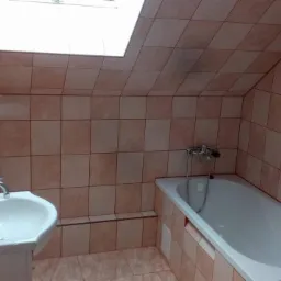 Ukończona łazienka na poddaszu z beżowymi płytkami, białą wanną i umywalką oraz grzejnikiem drabinkowym.