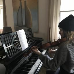 Kameralne.pl - Kobieta w czapce gra na skrzypcach, siedząc przy czarnym pianinie marki Bechstein, nuty na pulpicie, obraz z wazonami w tle.