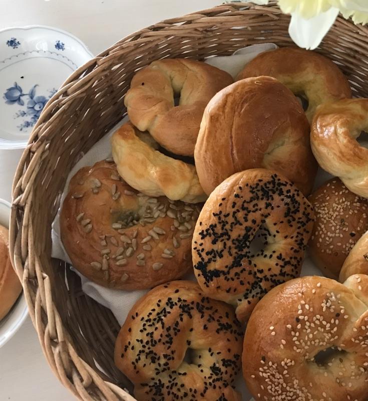 Kosz wiklinowy wypełniony różnorodnymi bajglami: z makiem, sezamem i słonecznikiem, na białym tle z kwiatami i talerzem w tle.