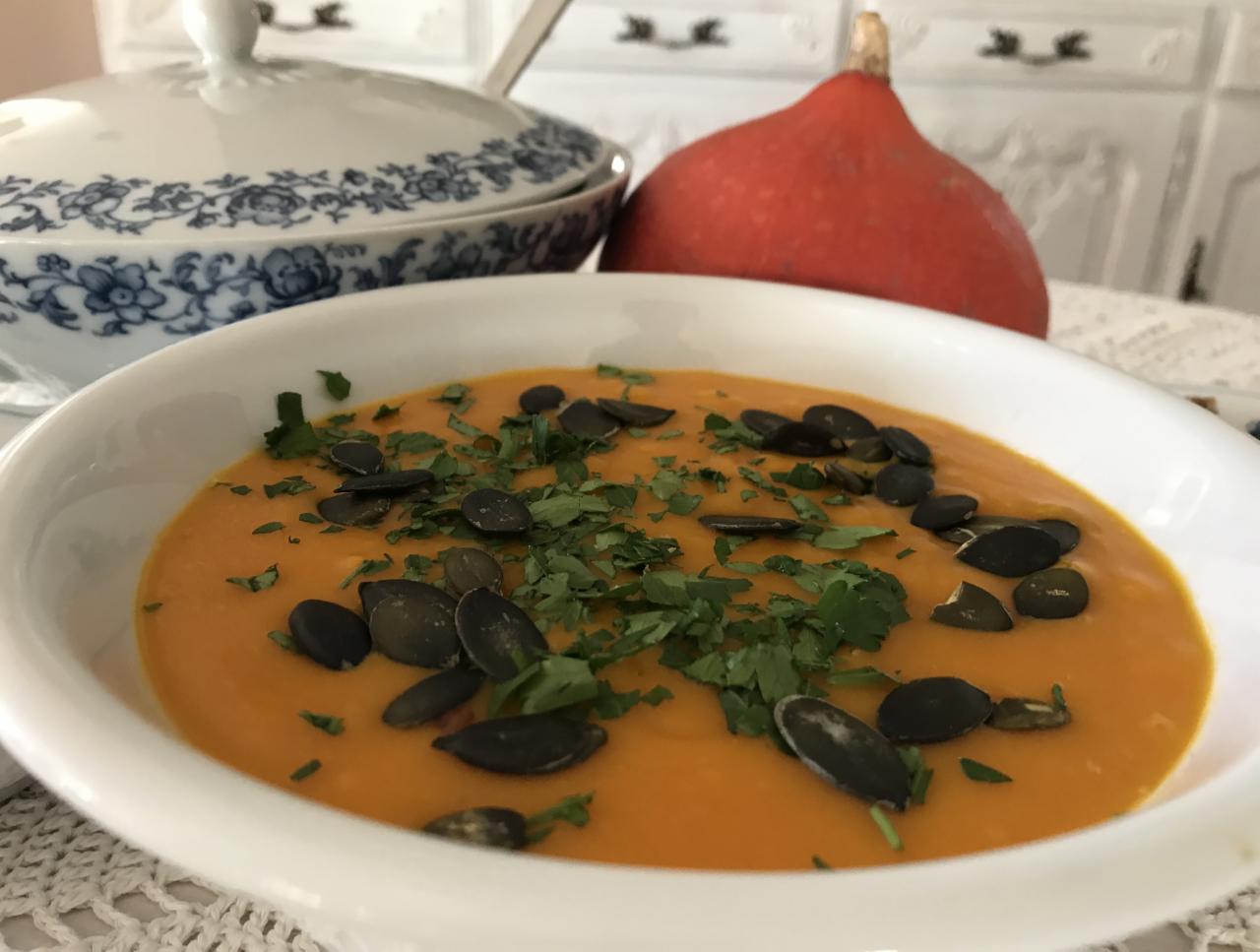 Krem z dyni hokkaido w białej miseczce, udekorowany pestkami dyni i świeżą kolendrą, z dynią hokkaido w tle i porcelanową wazą z niebieskim wzorem.