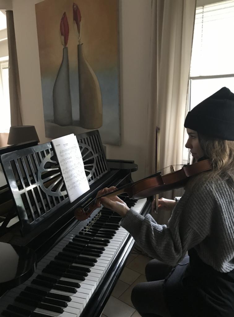 Kobieta w czapce gra na skrzypcach, siedząc przy czarnym pianinie marki Bechstein, nuty na pulpicie, obraz z wazonami w tle.