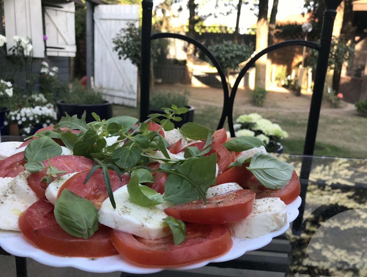 Caprese na białym talerzu, udekorowana świeżymi ziołami, serwowana na szklanym stole na tle ogrodu z białymi kwiatami i drewnianą altaną.