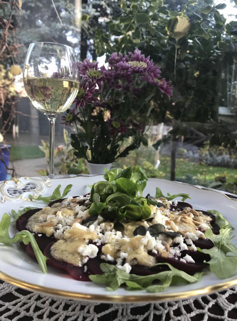 Elegancko podana sałatka z buraków z serem feta, sosem i rukolą na ozdobnym talerzu, w tle kieliszek białego wina i kwiaty.