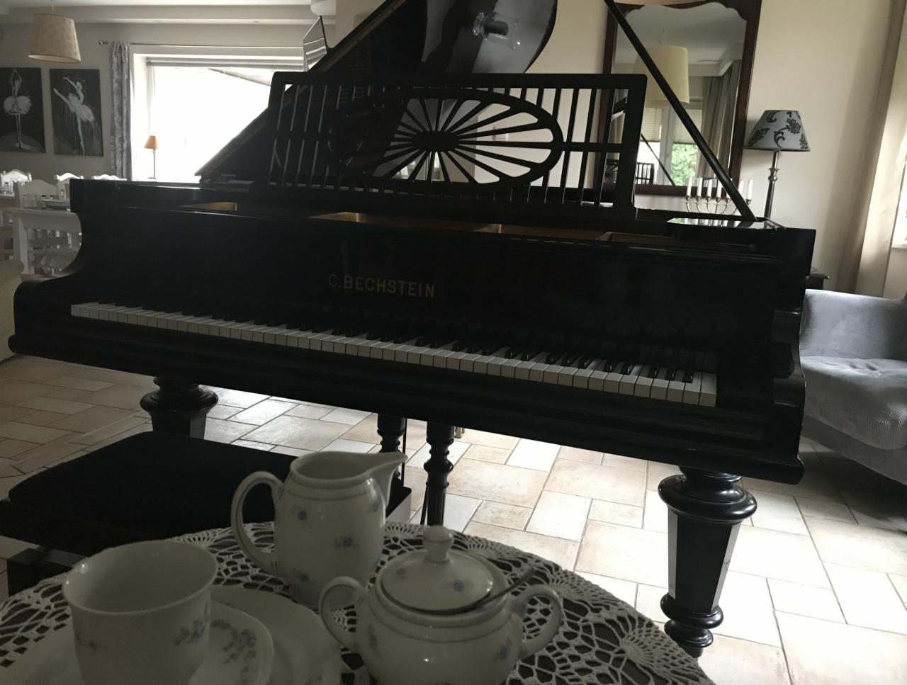 Czarny fortepian C. Bechstein w salonie z serwisem do herbaty na stoliku z koronkowym obrusem, w tle okno i obraz z baletnicami.