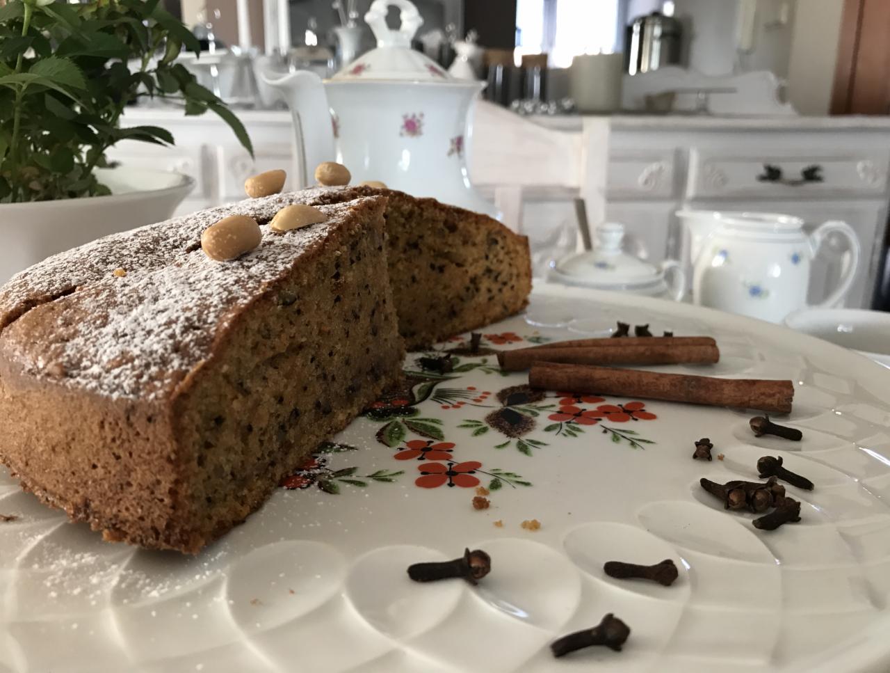 Kawałek ciasta posypany cukrem pudrem i orzechami na talerzu z motywem kwiatowym, obok laski cynamonu i goździki, w tle porcelanowa zastawa i biała komoda.