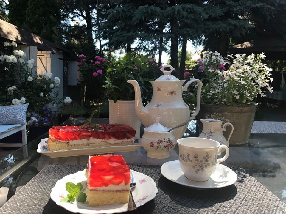 Letni stół w ogrodzie z ciastem truskawkowym w galaretce, porcelanową zastawą do herbaty, kwiatami w donicach i zielenią drzew w tle.