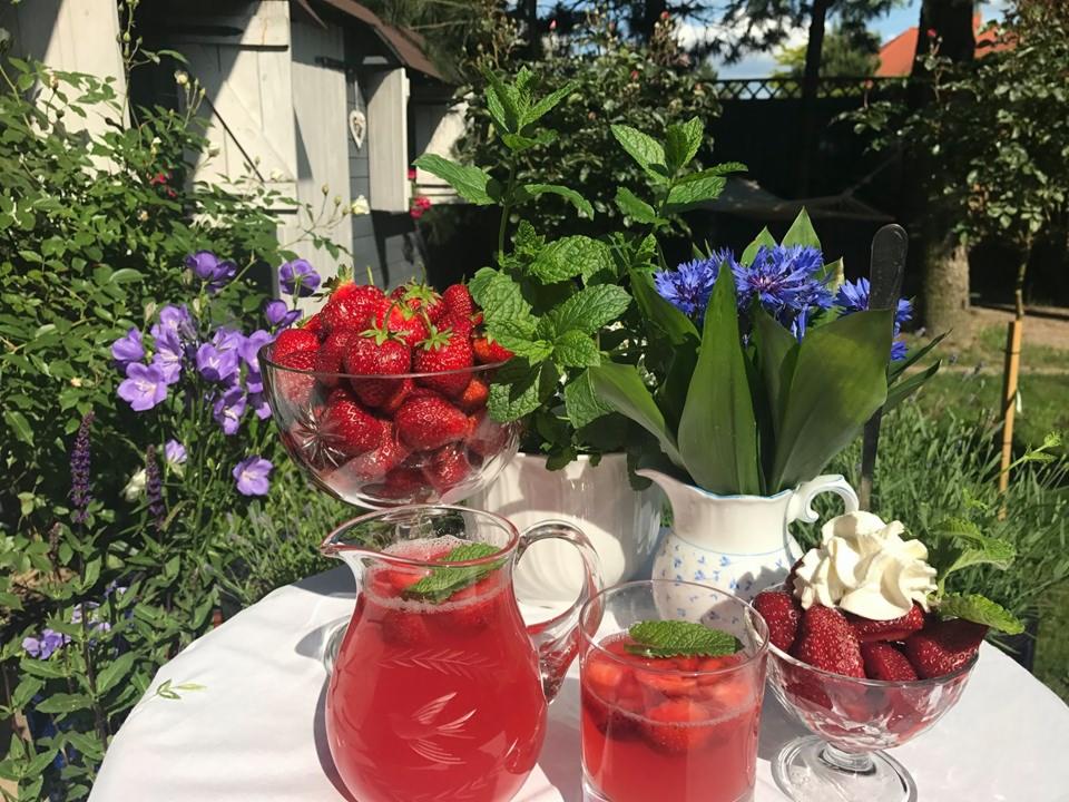 Letnia kompozycja na stole w ogrodzie: dzbanek i szklanka z napojem truskawkowym z miętą, miseczki truskawek ze śmietaną i bez, bukiet bławatków i mięty, w tle zielona roślinność i drewniany domek.