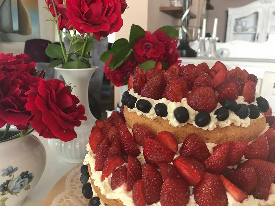Dwupoziomowy tort z bitą śmietaną, truskawkami i borówkami, udekorowany czerwonymi różami w wazonach na stole.